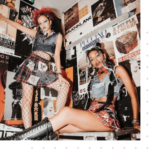 Two girls sitting in a room covered in posters sitting and standing in matching alternative fashion